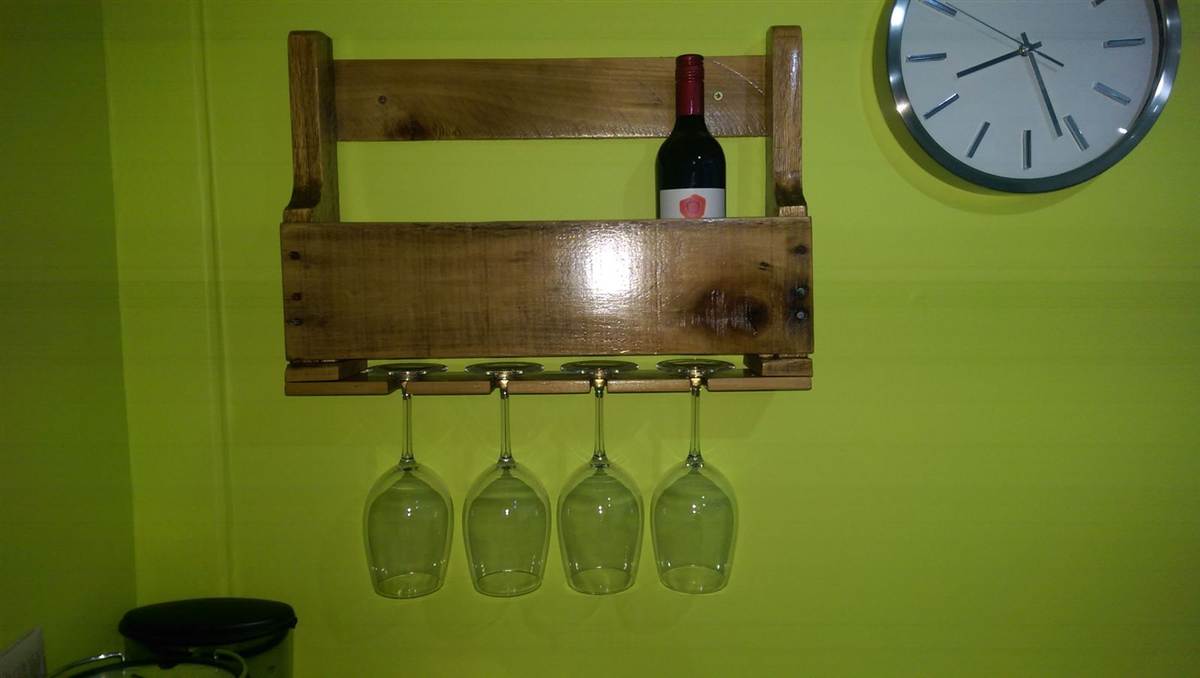Finished wine rack mounted on a wall — oiled pallet wood, bottle shelf on top, four wine glasses hanging inverted below